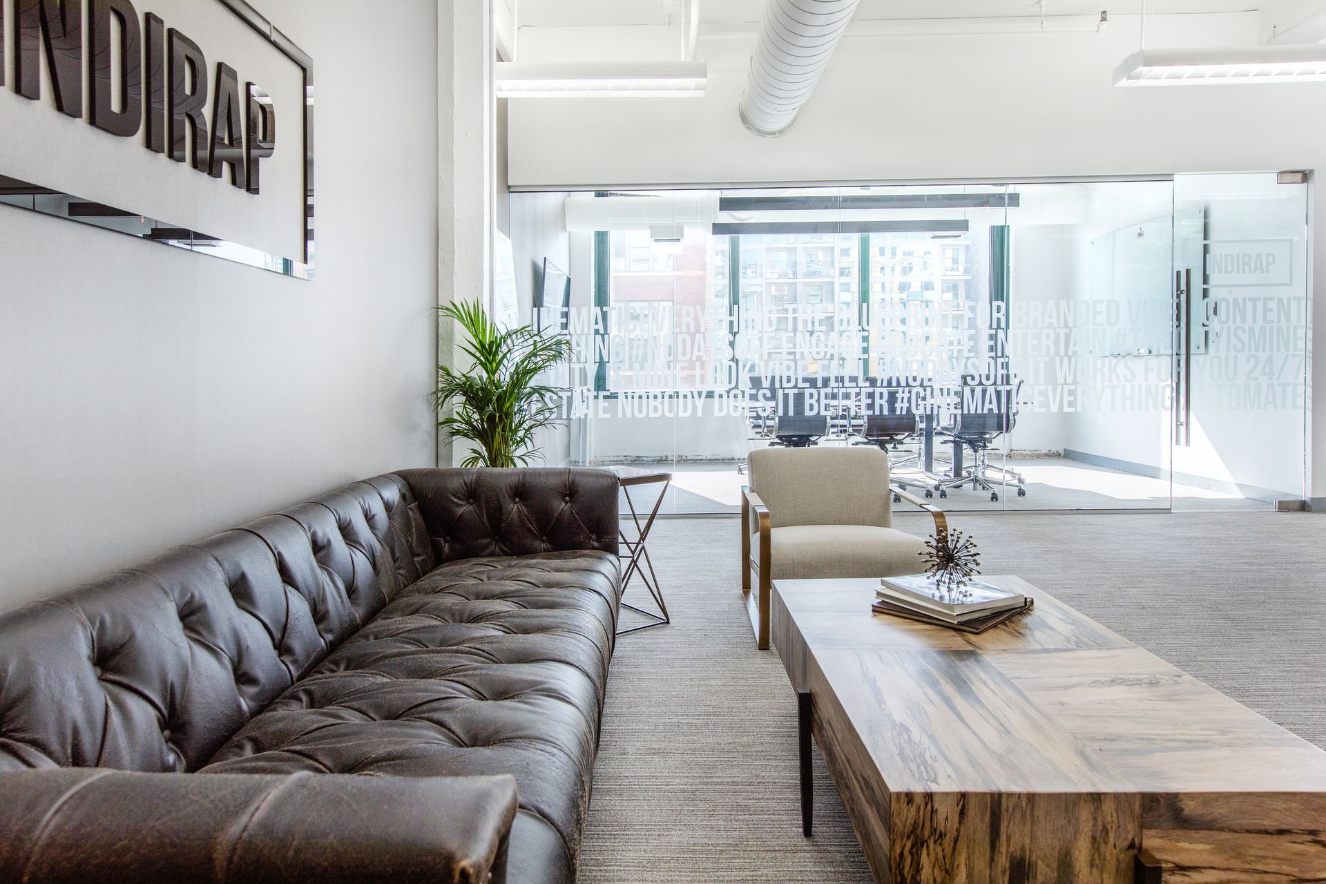 Leather sofa in a modern office waiting area; coffee table, arm chair, glass wall with meeting room visible.