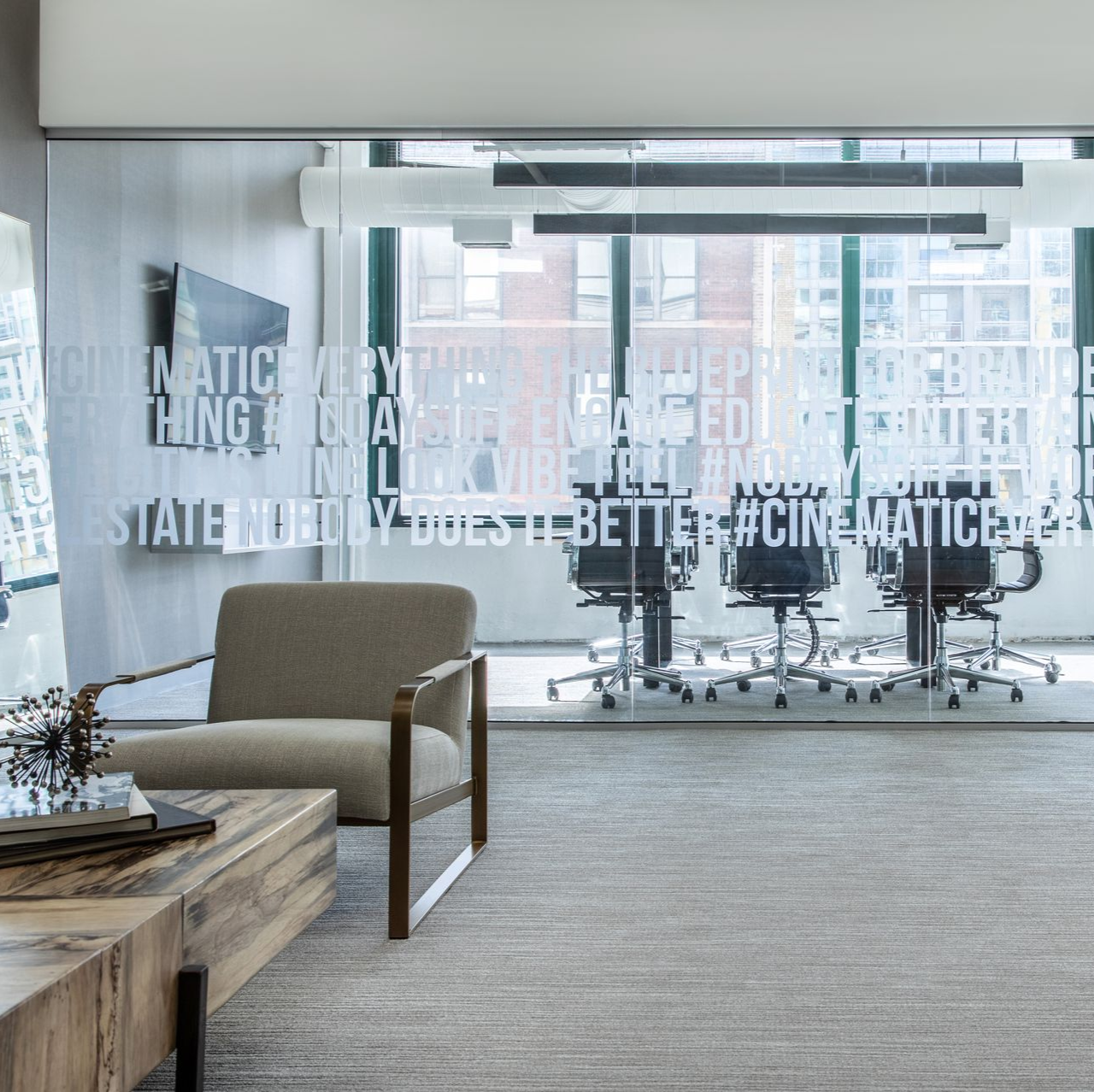 Modern office waiting area with a chair, coffee table, and conference room visible through a glass wall.