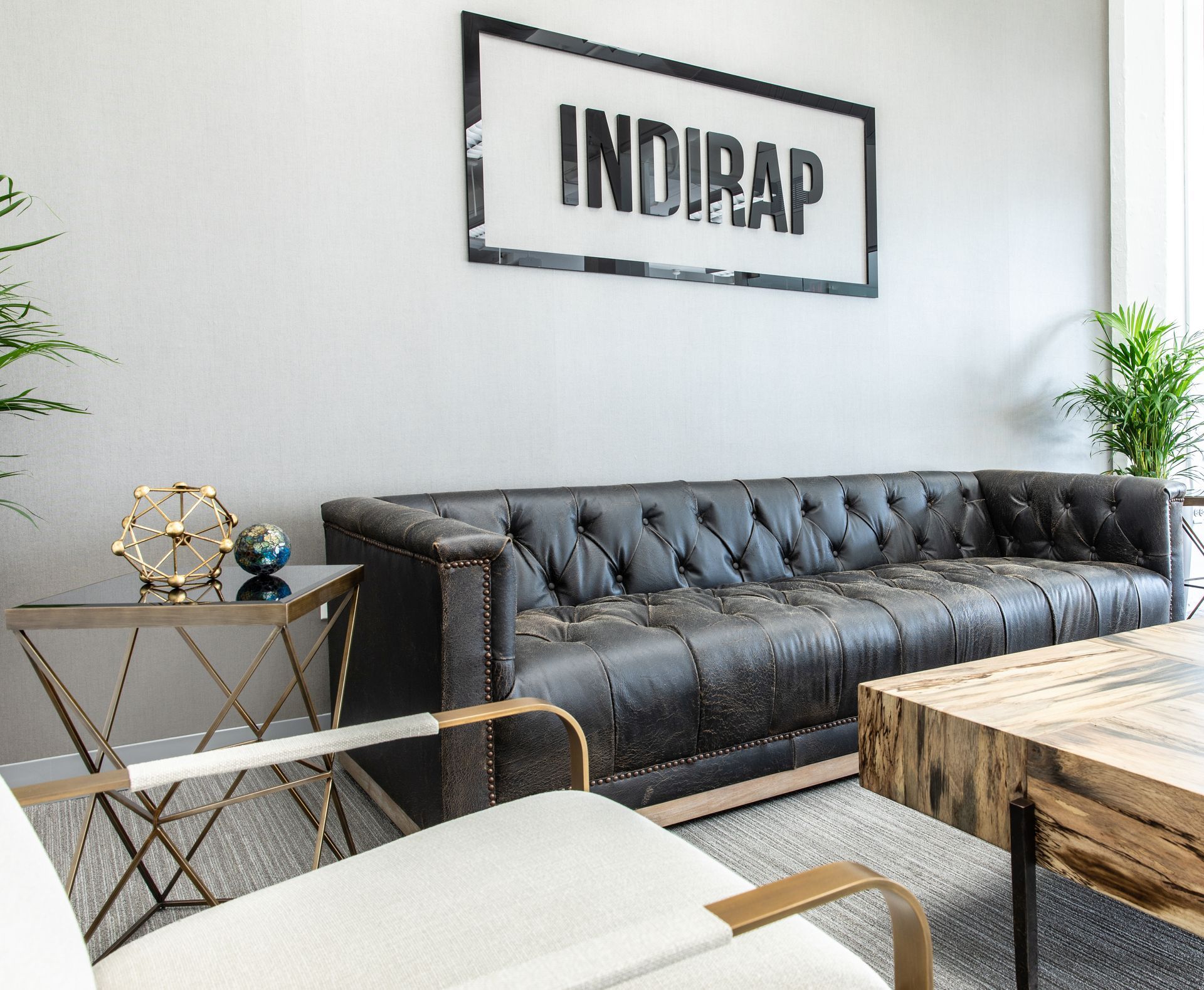 Lounge area with black leather sofa, modern chair, and wooden coffee table. Sign: INDIRAP. Gray walls, carpet.