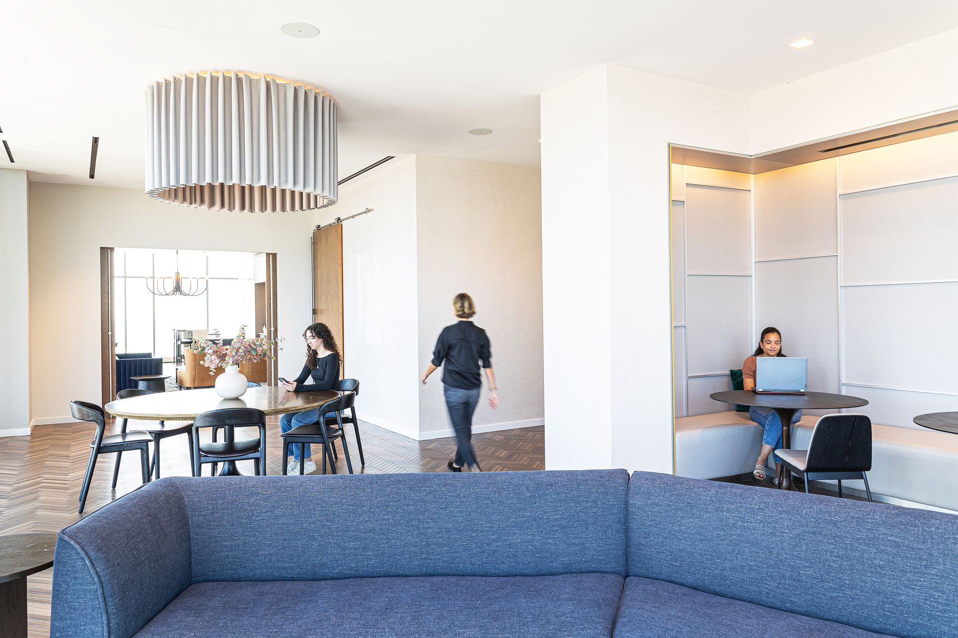 Modern office space with people working at tables, a blue sectional couch, and a large light fixture.