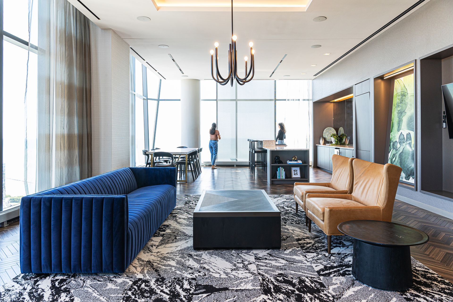 Modern living room with blue sofa, leather chairs, and large windows. People stand near a dining table and counter.