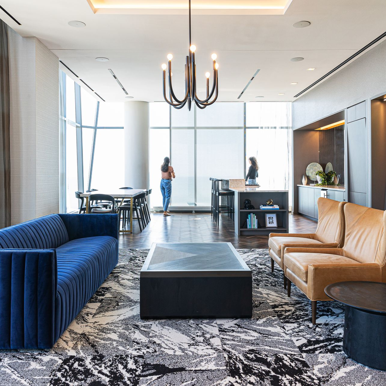 Modern hotel lobby with blue velvet sofa, tan chairs, patterned rug, chandelier, and floor-to-ceiling windows