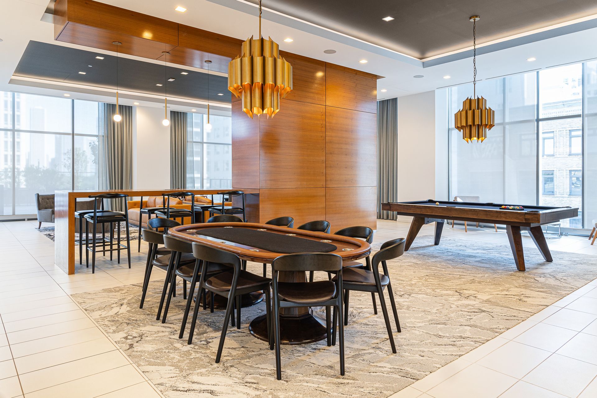 A modern lounge area with a poker table, pool table, bar stools, and two gold pendant lights.