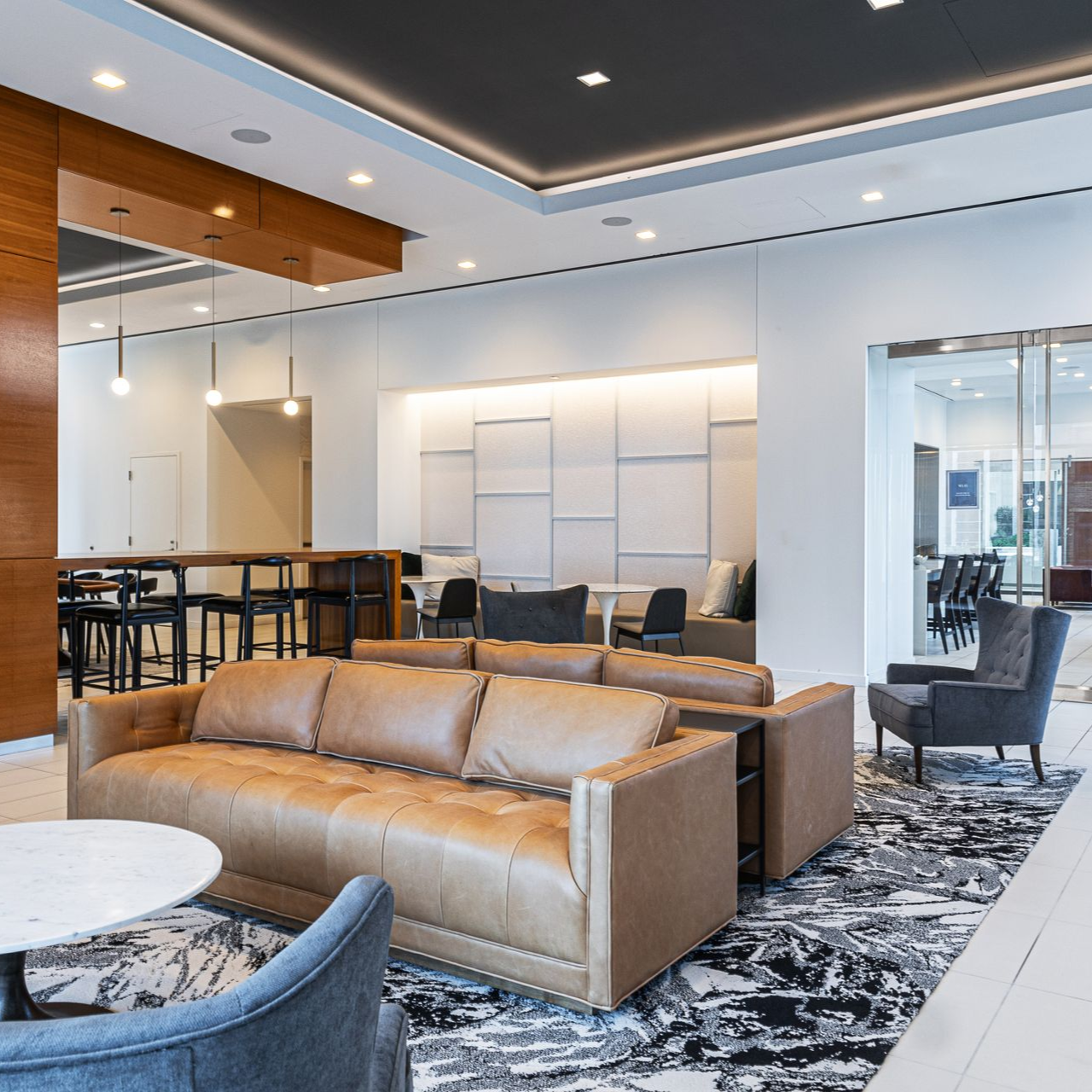 Lobby with brown leather sofa, chairs, marble table, rug, and bar seating. White walls, wood accents, and recessed lighting.