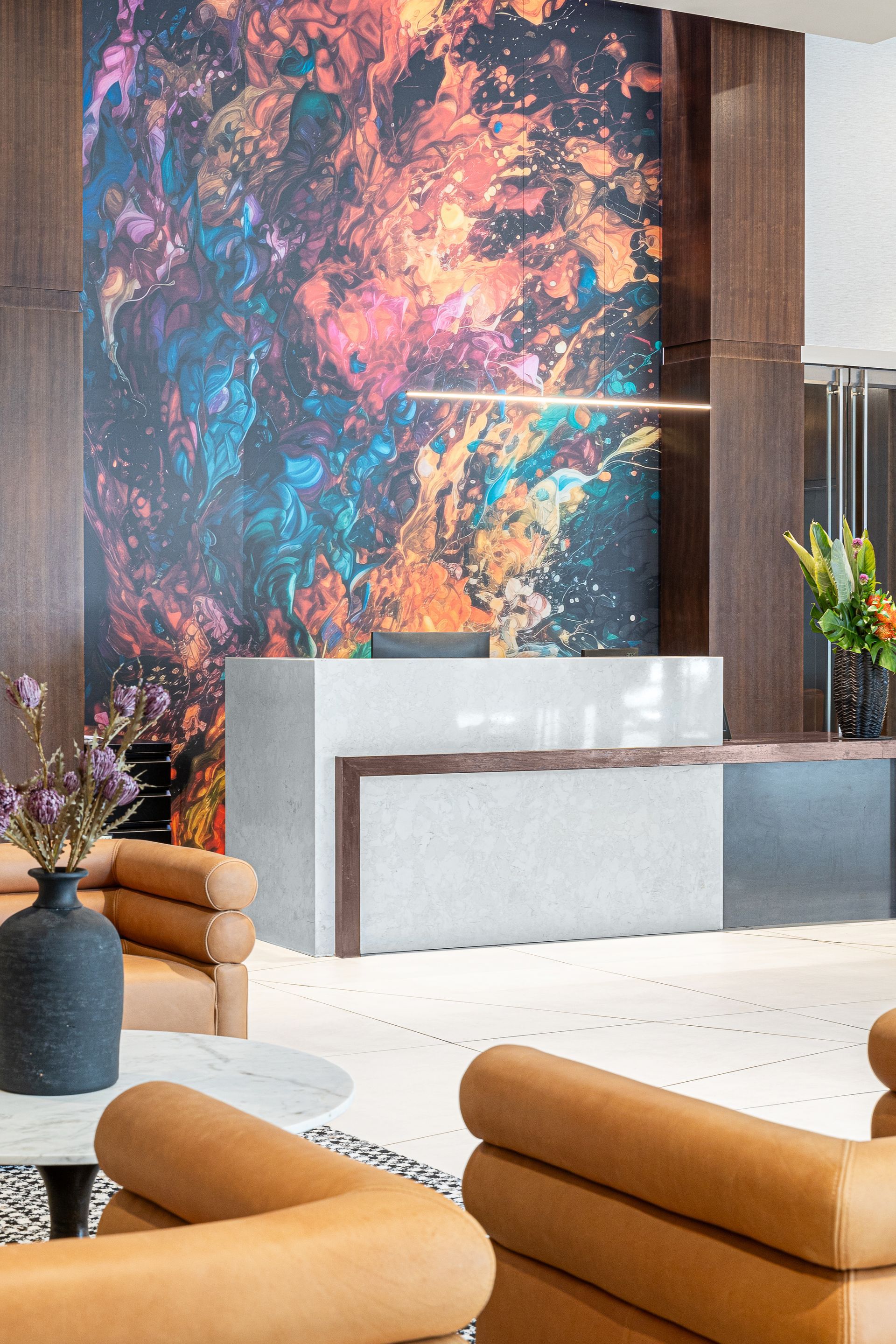 Reception area with abstract art backdrop, modern desk, tan chairs, and floral arrangements.