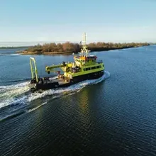 Yellow workboat with equipment on blue water near a tree-lined shoreline