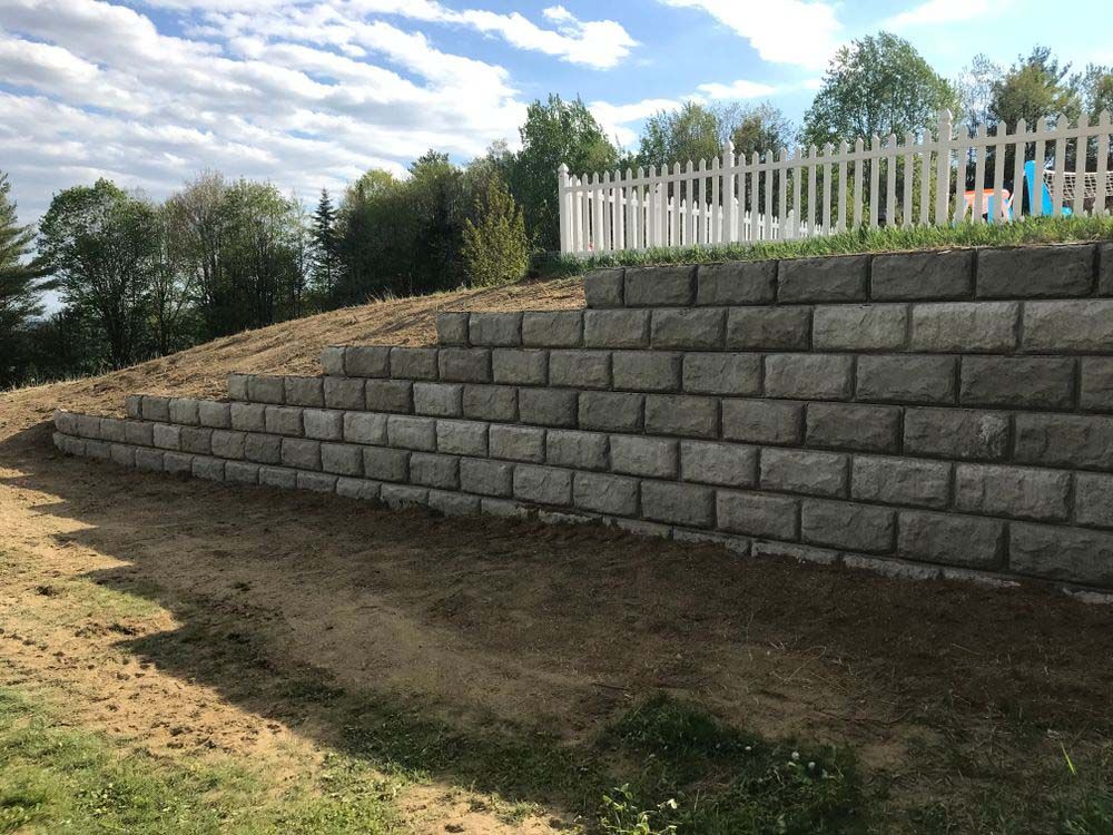 A large brick wall is sitting on top of a dirt hill next to a white picket fence.