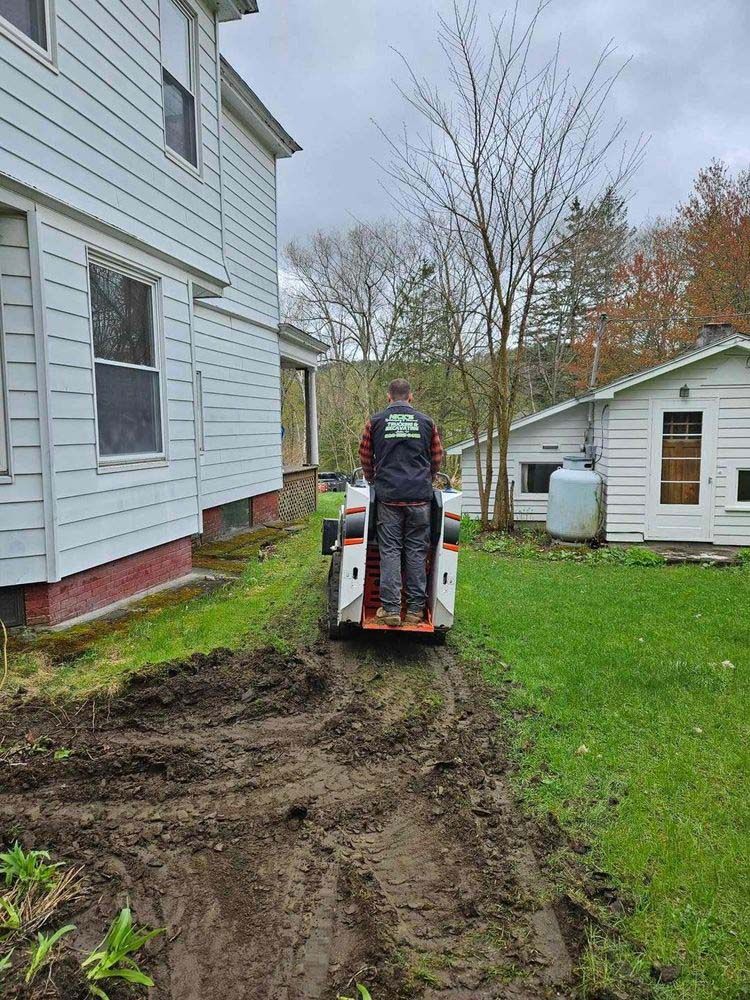 A man is riding a tractor down a dirt road in front of a house.