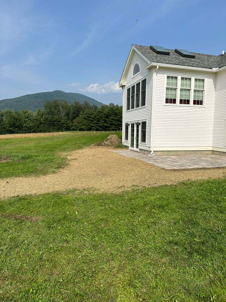 A white house is sitting in the middle of a grassy field with mountains in the background.