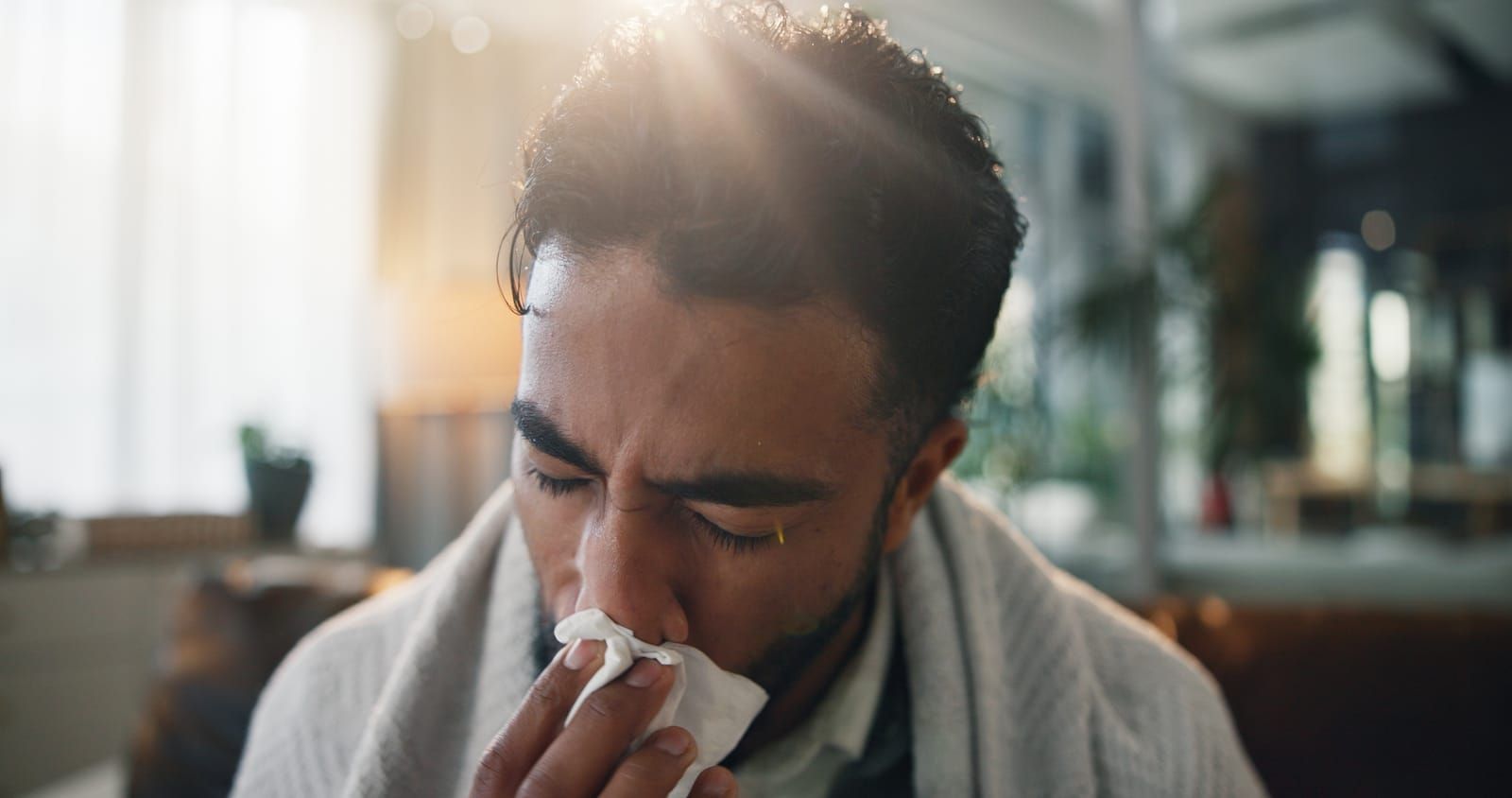 Man blowing his nose with a tissue, wrapped in a blanket, indoors.