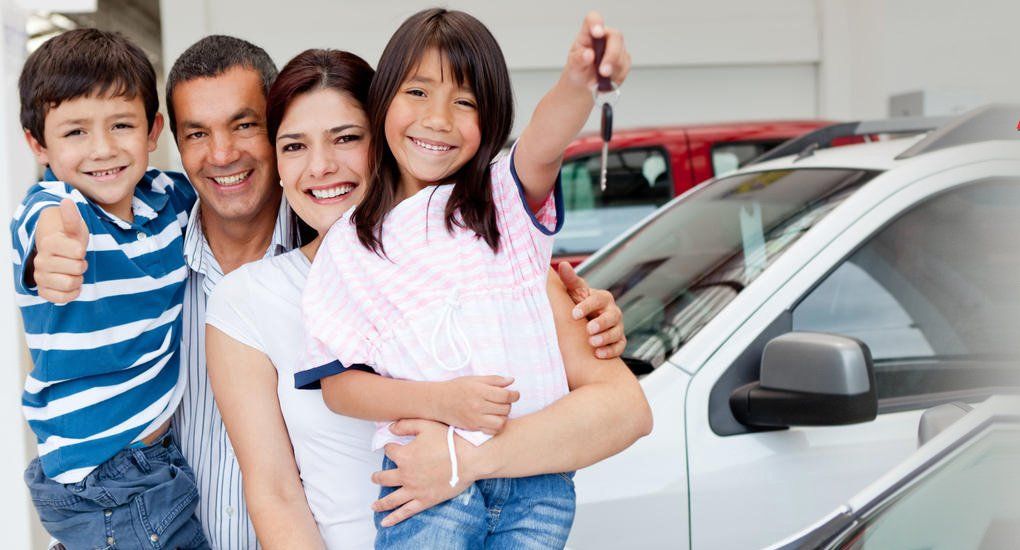 Family of four smiling, holding car keys next to a new white car at a dealership.