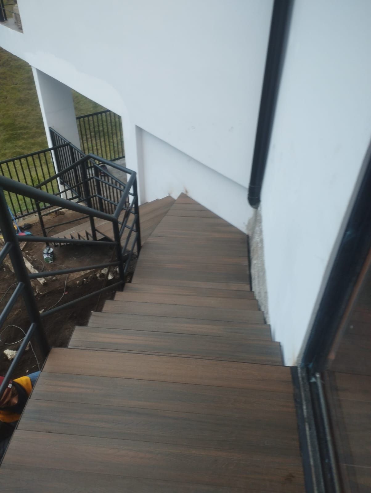 Escalera de madera que desciende junto a un edificio blanco con barandillas negras y una ventana.