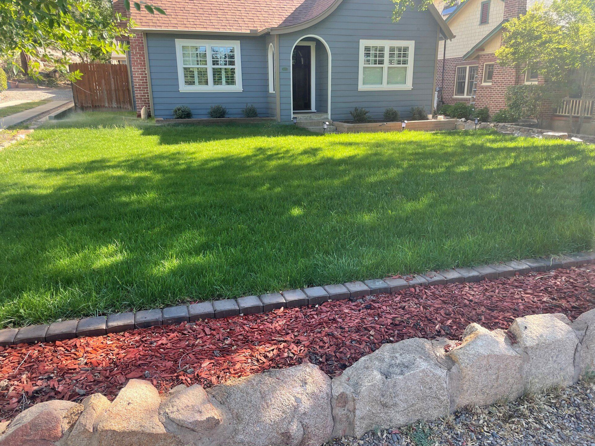 Blue house with green lawn, red mulch bed, and rock edging under a sunny sky.