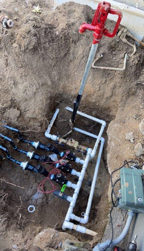 Excavated ground with irrigation system and red water pump. White PVC pipes, black valves, and green timer box visible.