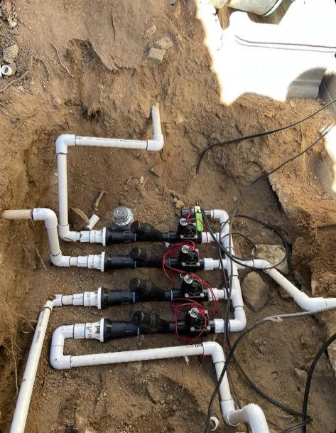 Irrigation system in a dirt trench, with PVC pipes, valves, and wiring, outdoors.