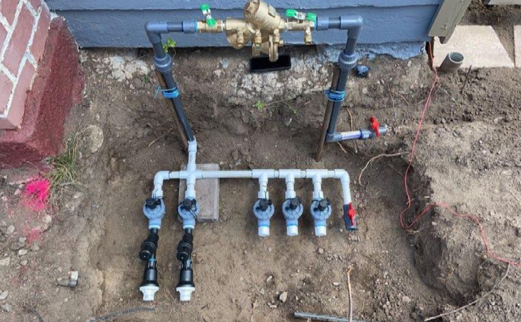 Irrigation system with PVC pipes, valves, and a water meter installed in a dirt ground near a house exterior.