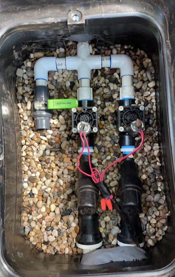 Irrigation system valves in a black plastic box filled with rocks. White PVC pipes and black solenoid valves.