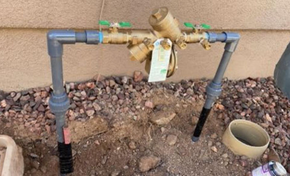 Water pipe assembly with valves and a meter, in an outdoor setting with gravel.