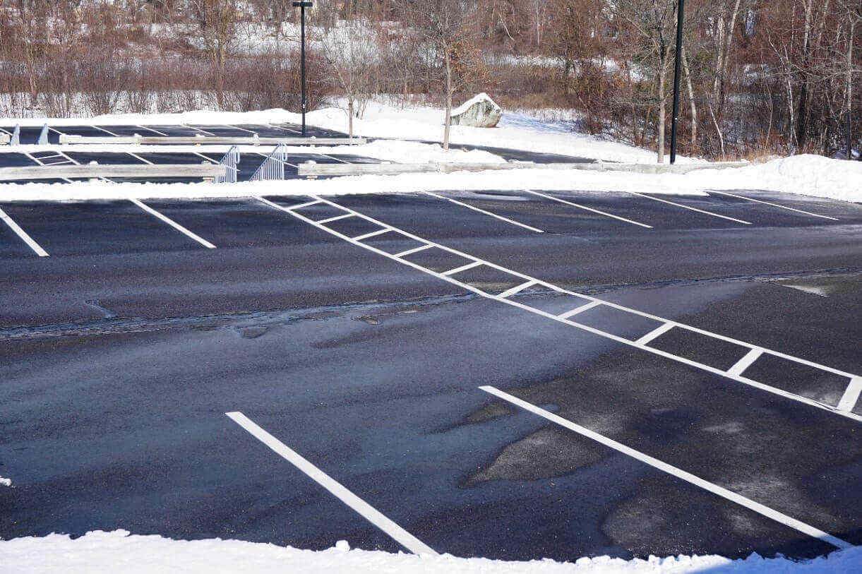 Snowy parking lot with white lines, partially cleared, with trees in the background.