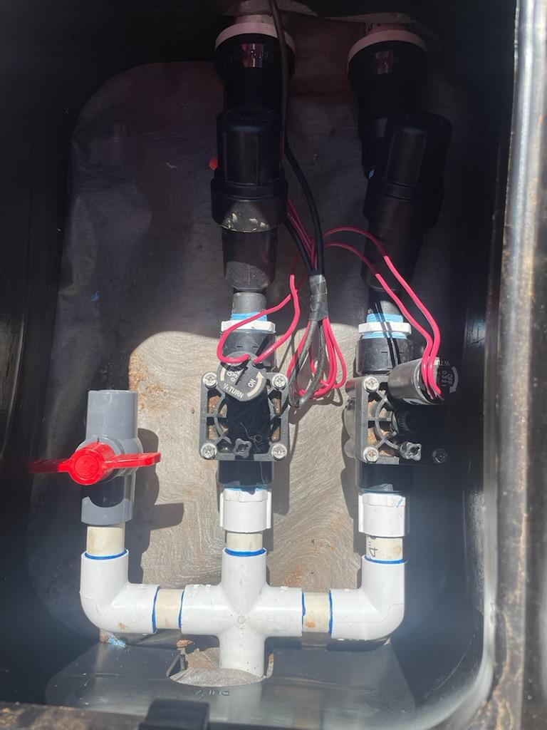 Irrigation valve box with two valves, PVC pipes, and electrical wiring; includes a red-handled shutoff valve.