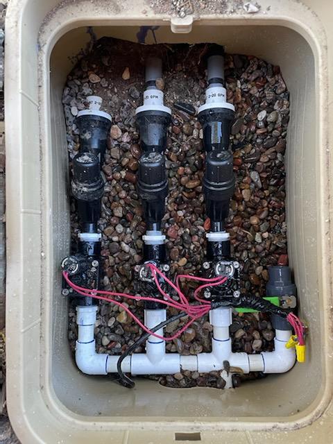 Irrigation system with three black valves and white PVC pipes in a beige plastic box filled with gravel.