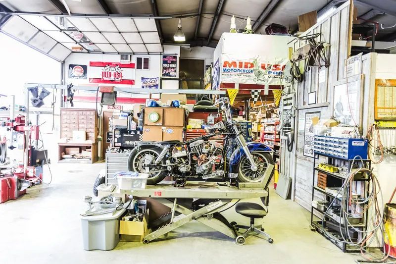 Motorcycle on a lift in a cluttered workshop with tools, shelves, and various signage.
