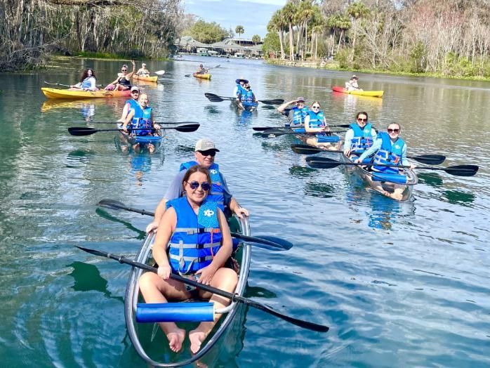 People kayaking in clear kayaks on a river, wearing life vests. Trees line the banks, sunny day.