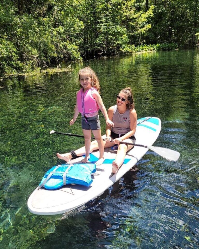 Young girl and woman paddleboarding on clear water in a sunny, wooded area.