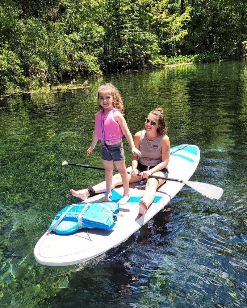 Woman and child paddleboarding on clear water, sunny day. Child in a life vest smiles.