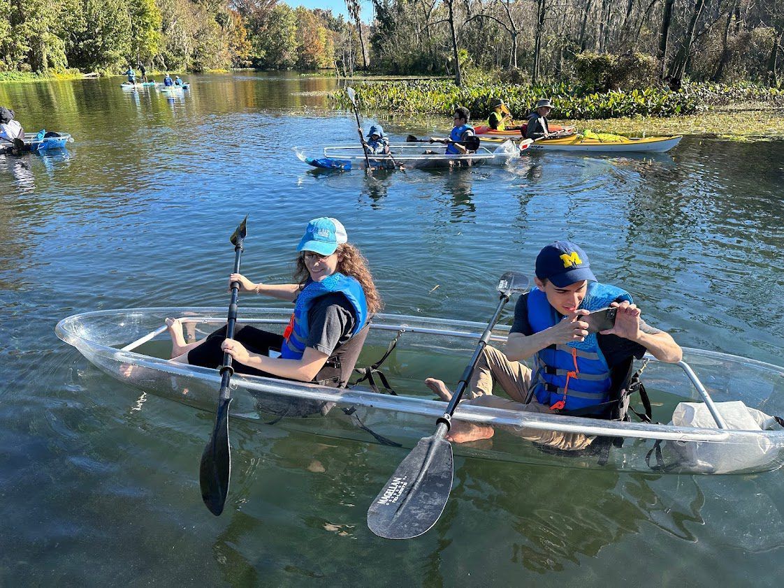 Two people kayaking in a clear boat on a lake with others in the background, paddling on a sunny day.