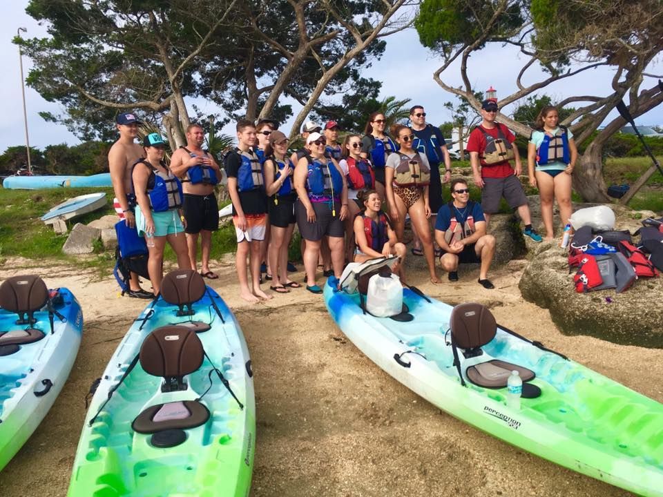 Group of people by kayaks on a beach, wearing life vests.