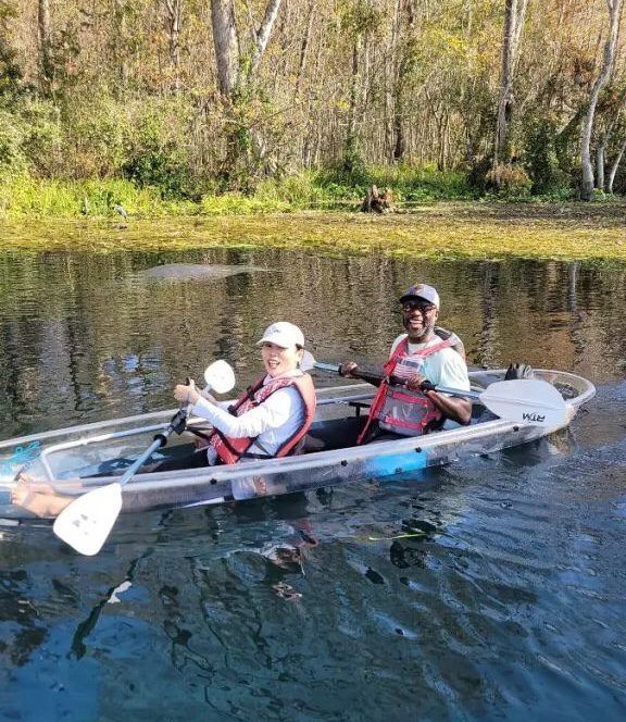 Two people kayaking in a clear kayak on a river. One gives a thumbs up. Sunlight reflects on the water.