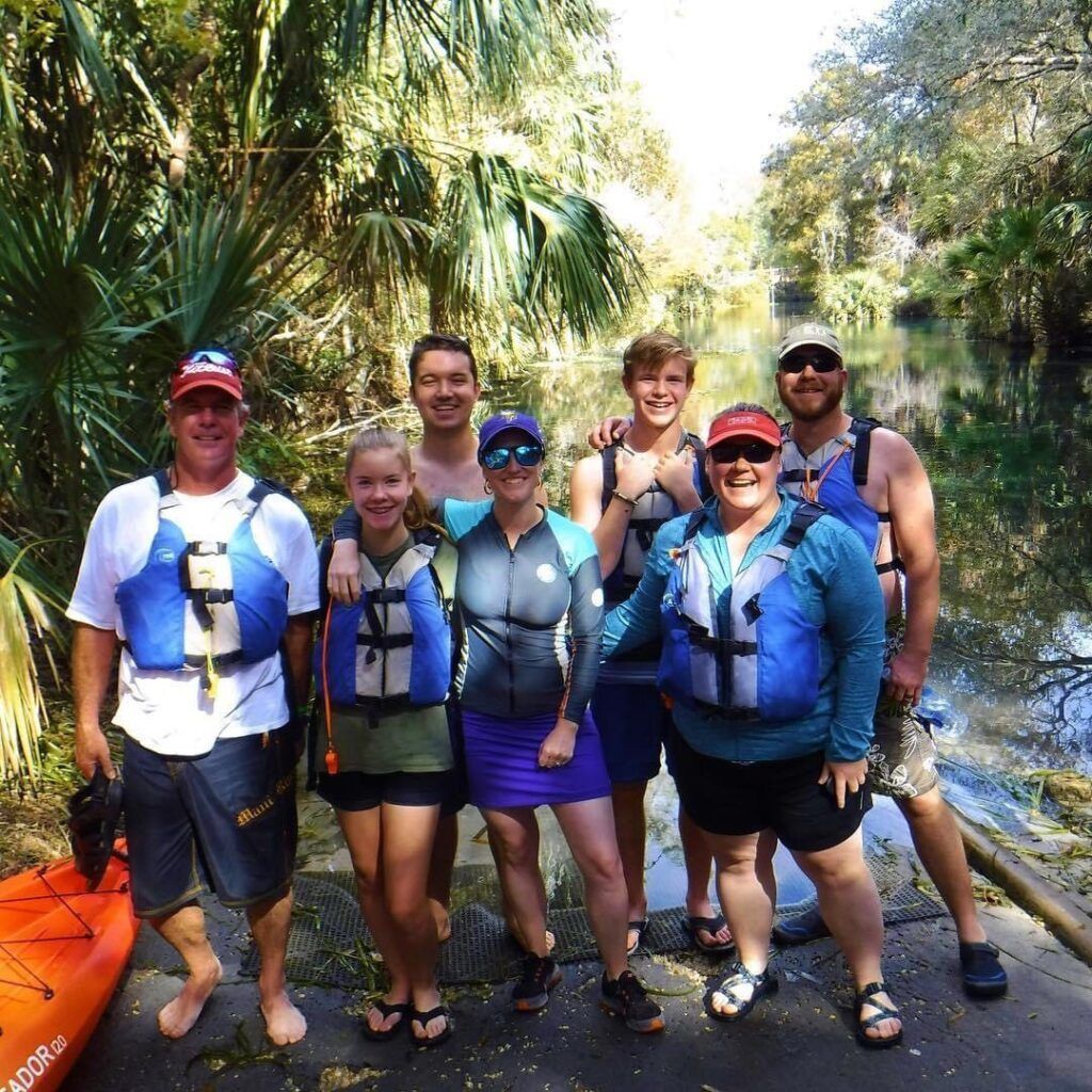 Group of people in life vests pose near a kayak and waterway, surrounded by greenery.