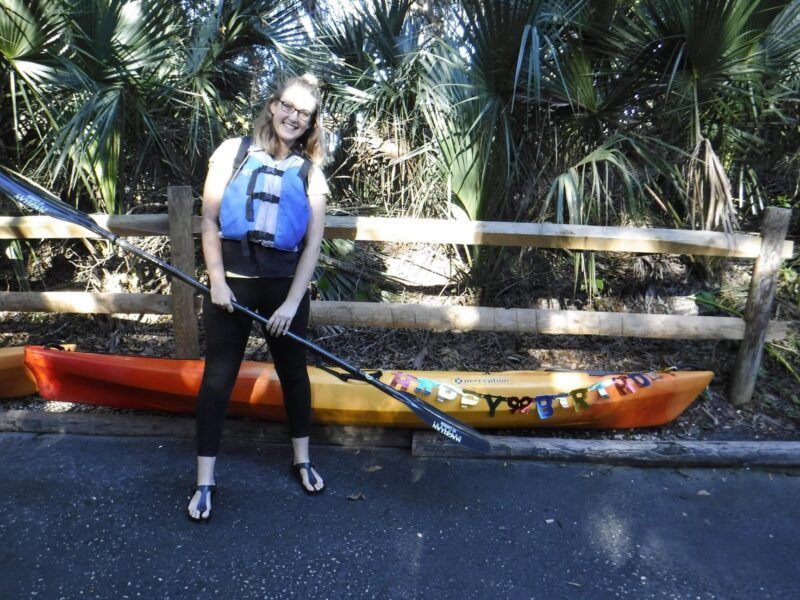 Woman holding a paddle, wearing a life vest, next to kayaks and a wooden fence, smiling outdoors.