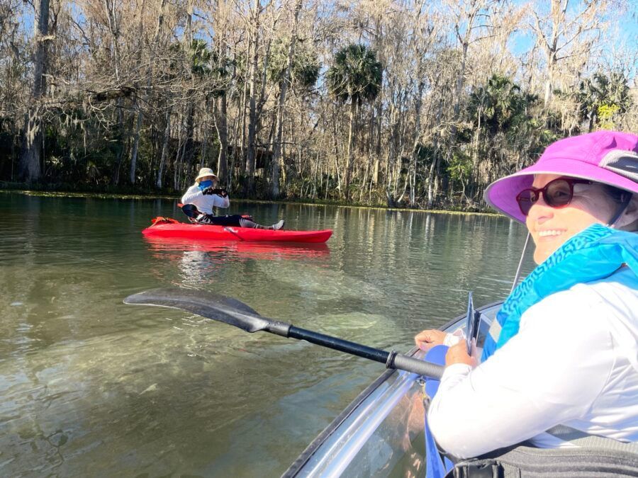 Person kayaking on a river with another person in a red kayak in the background. River scene with trees and blue sky.