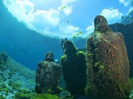Underwater statues covered in algae, facing upward, with sunlight filtering through the water.