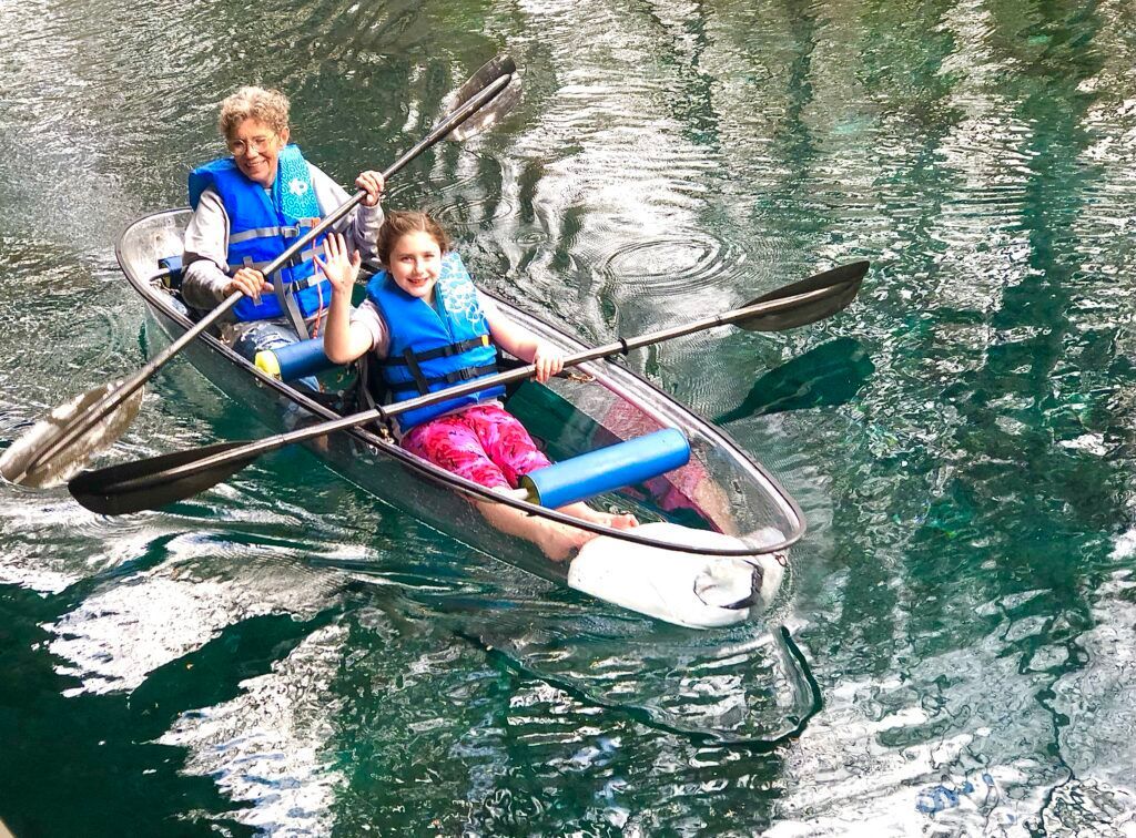 Two people kayaking in a clear kayak, waving on a lake.