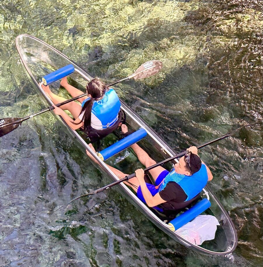 Two people in a clear kayak, paddling on a clear river. Both wear life vests.