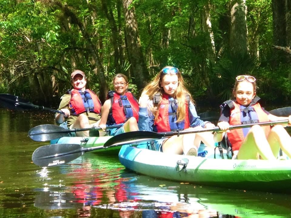 Four people kayaking on a river under a canopy of green trees. They wear life vests and hold paddles.