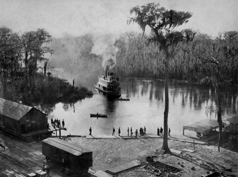 Steamboat on river approaches dock, people gather; trees line the banks.