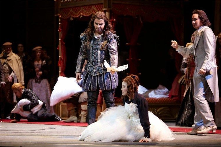 Opera scene: Actors in Renaissance attire. Man in blue stands over kneeling woman in white gown.