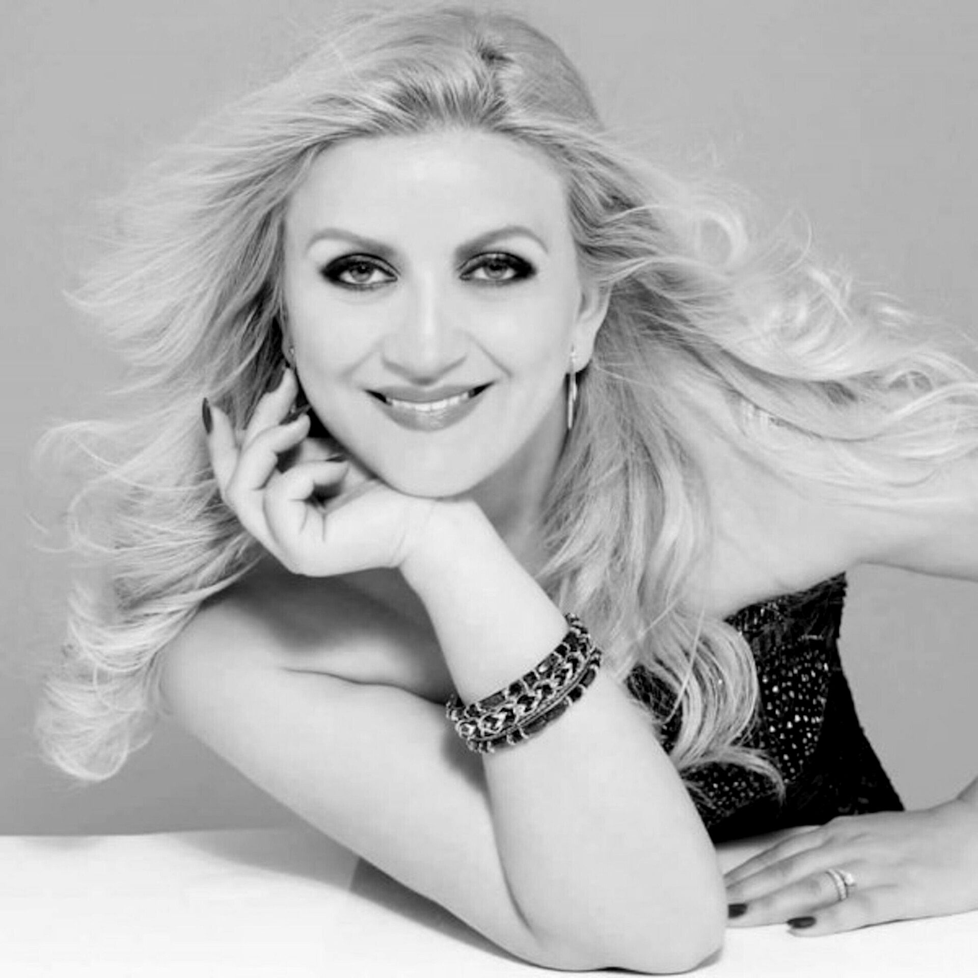 Blond woman smiling, resting chin on hand, wearing a bracelet and a strapless top.