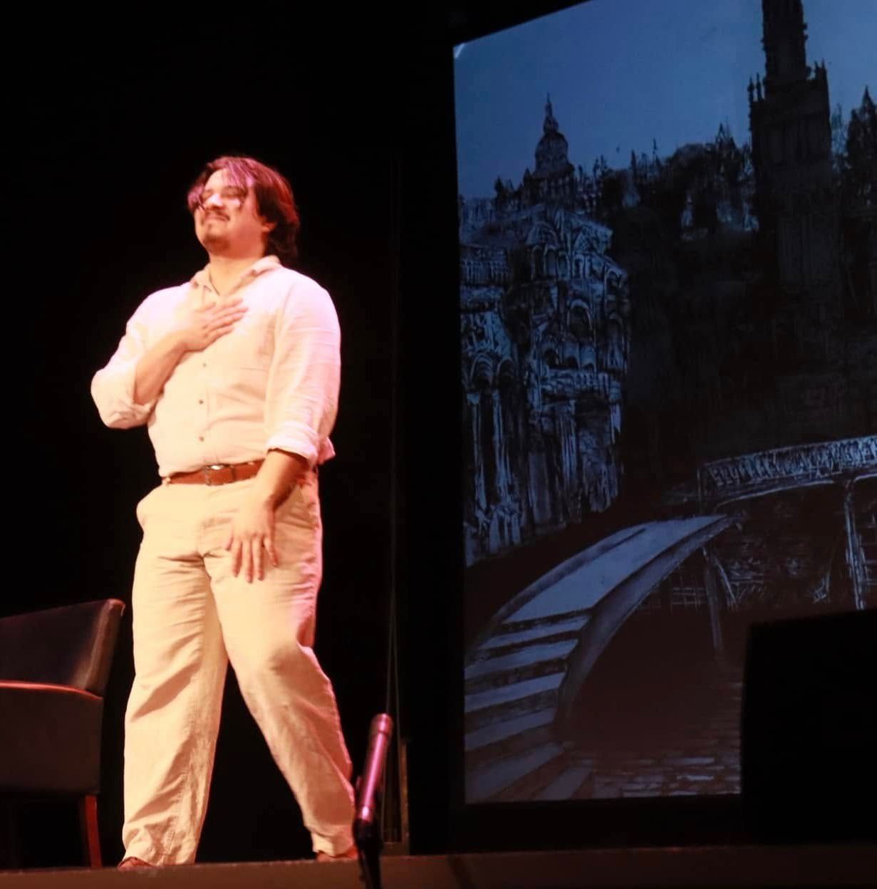 Man on stage with hand on chest, gesturing toward a castle image. He's wearing white and tan clothing.
