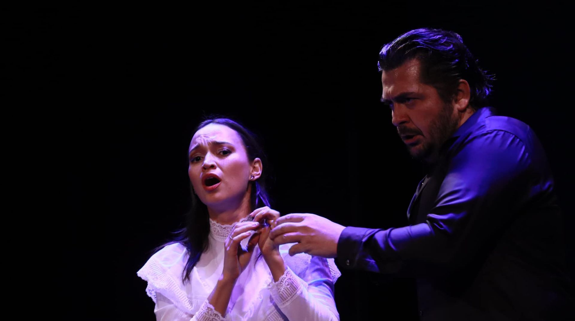 A woman in white and a man in black on stage singing, dramatic expressions.