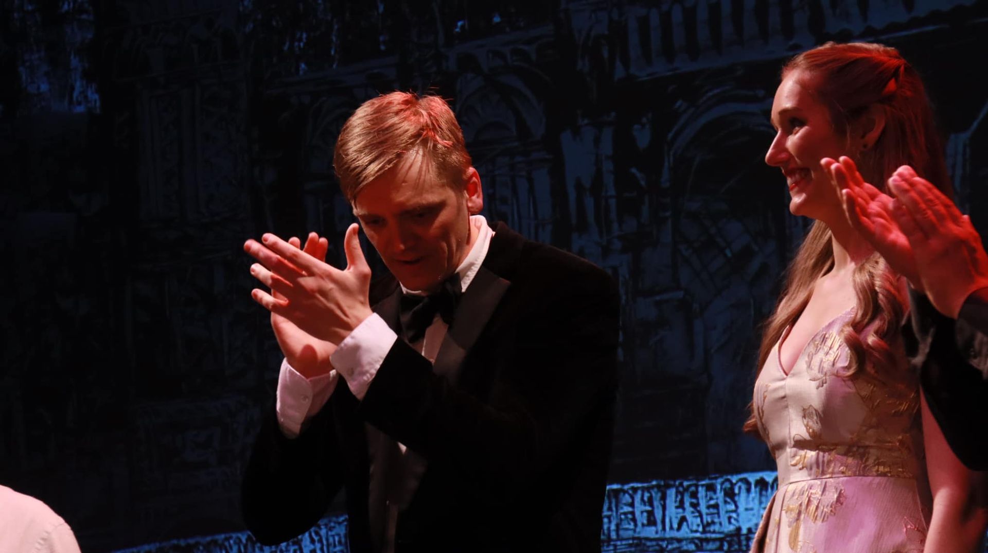 Man in tuxedo applauds onstage; woman in gown smiles, also applauding. Stage background: building silhouette.