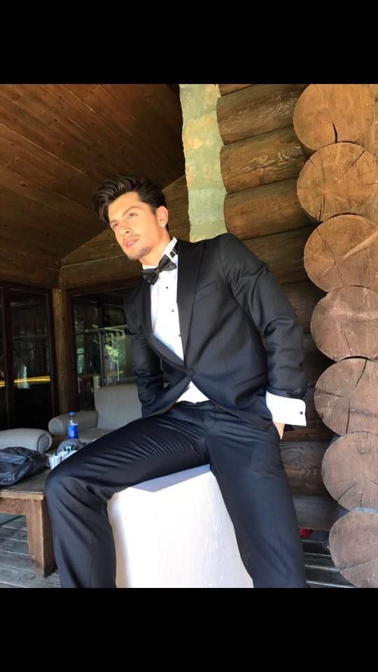 Man in black tuxedo poses outside, leaning against a log cabin wall.