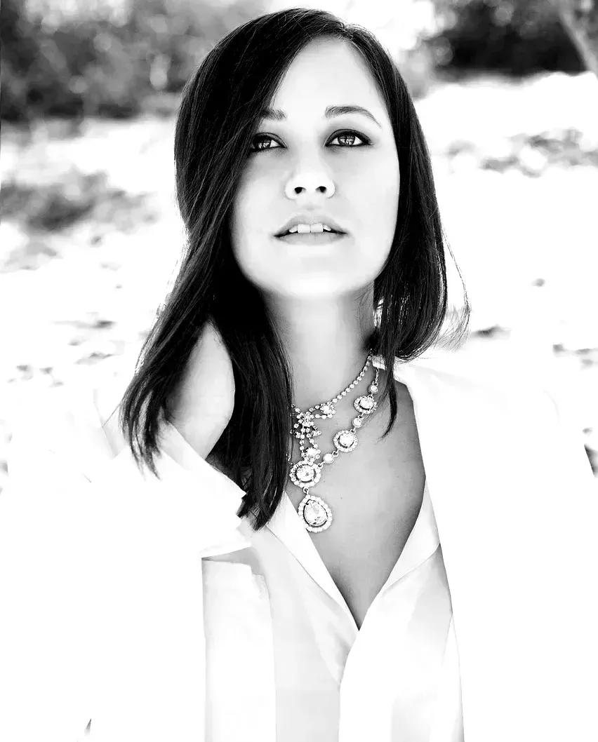 Woman with dark hair, wearing a white shirt and necklace, looking at the camera.