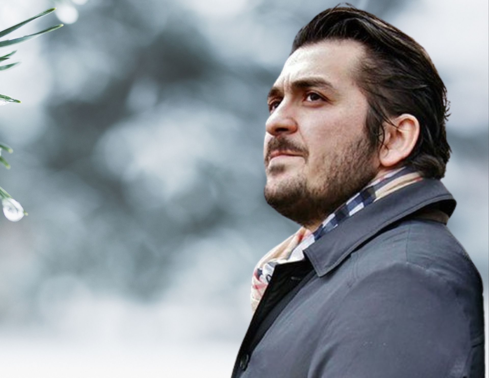 Man in a coat and scarf looking upward, outdoors with a blurred, wintery backdrop.
