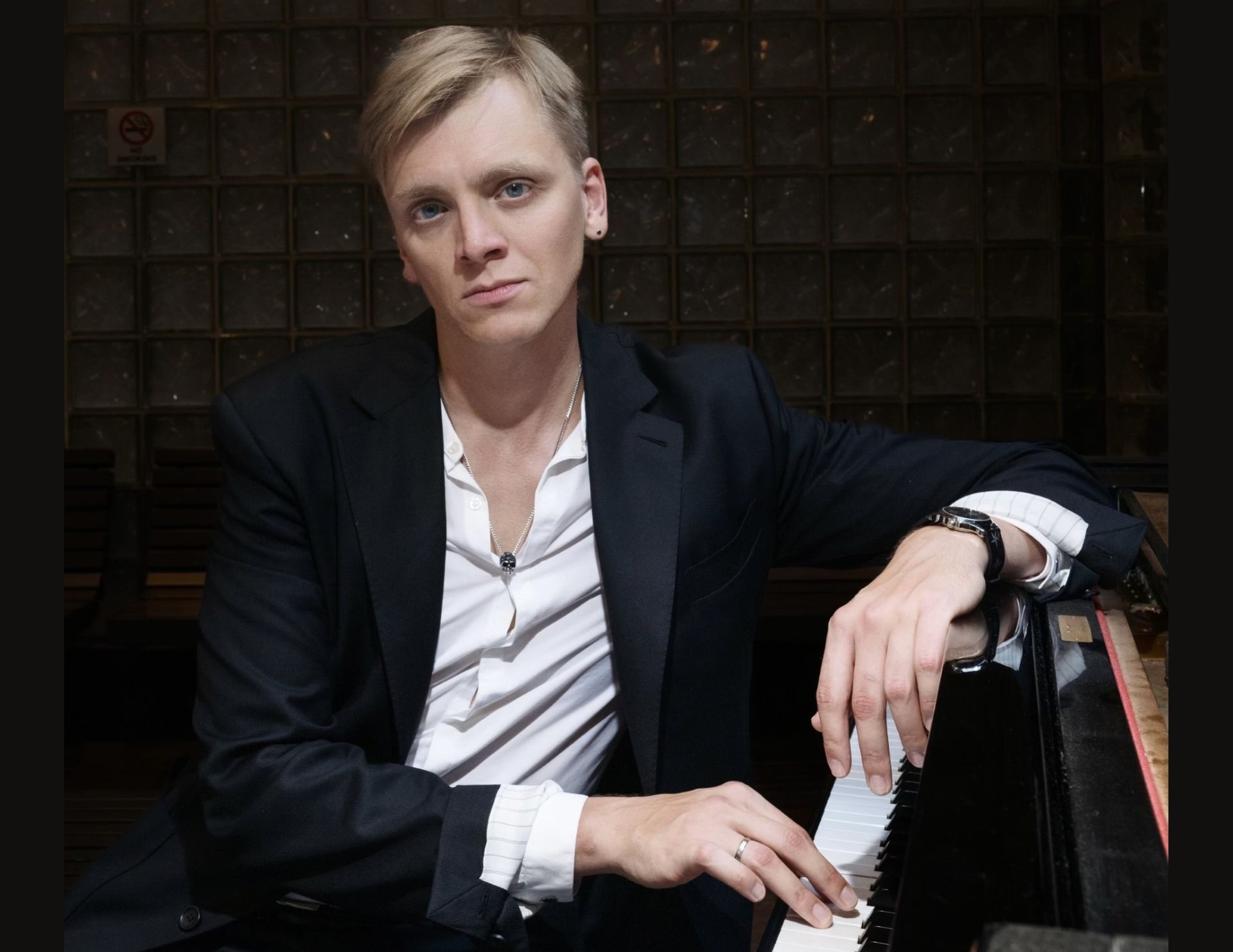 Blond man in blazer, leaning on piano, looking at camera, serious expression, dark background.