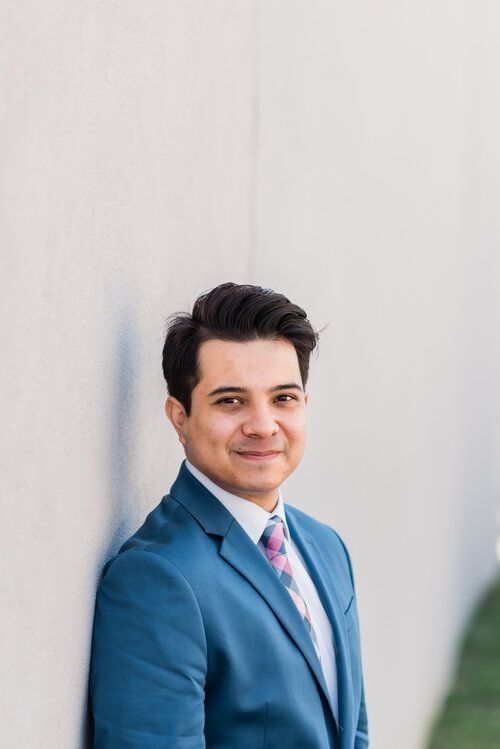 Man in blue suit, leaning against a light-colored wall, smiling.